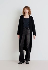 Cardigan lung negru peste un top închis la culoare și blugi negri evazați. Material texturat, siluetă fluidă și bocanci negri joși. Design simplu.