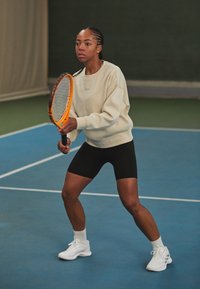 Tennisemängija kreemivärvilises sviitrises ja mustades lühikestes pükstes hoiab oranži reketit. Ta seisab sinisel väljakul rohelise taustaga.