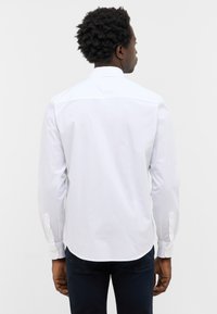 Biała długa koszula z długim rękawem, klasycznym kołnierzykiem, zapinana na guziki przy mankietach i z zaokrąglonym dołem. Gładki materiał bez wyraźnych wzorów czy faktur.