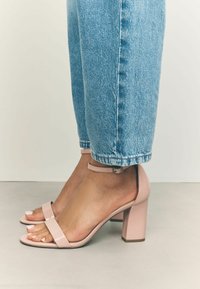 Sandali con tacco alto in pelle patent rosa, dotati di cinturino alla caviglia, tacco quadrato e punta aperta, abbinati a jeans azzurri arrotolati.