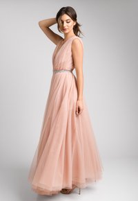 Różowa suknia z tiulu w kolorze brzoskwiniowym z głębokim dekoltem w kształcie litery V, zwiewną spódnicą i srebrnym zdobionym paskiem w talii. Gładki materiał o warstwowej teksturze.