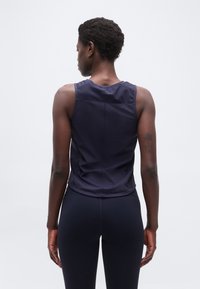 Γυναίκα πο υ φαίνεται από πίσω, φοράει ναυτικό μπλε αμάνικο αθλητικό τοπ και αντίστοιχο κολάν, με φόντο απλό φωτεινό.