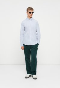 Blau-weiß gestreiftes Hemd mit Knöpfen, grüne Jogginghose, weiße und graue Turnschuhe. Person trägt Sonnenbrille und steht vor einem neutralen Hintergrund.
