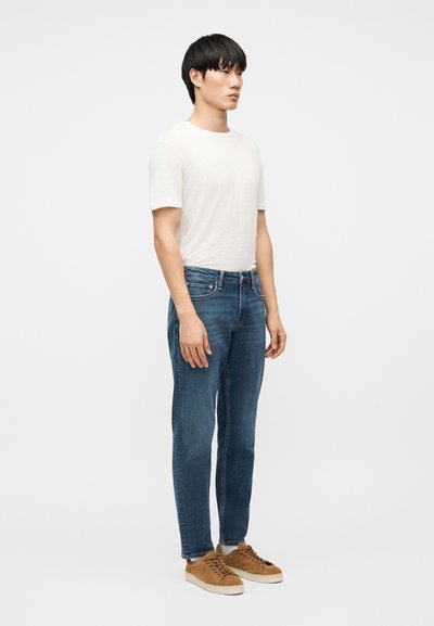 Homme debout portant une chemise blanche unie à manches courtes, un jean en denim bleu, des chaussettes blanches et des baskets en suède beige, sur un fond blanc.