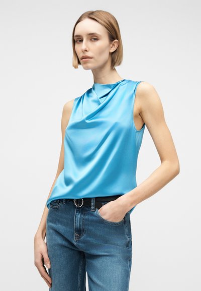 Γυναίκα με κοντά ξανθά μαλλιά, φορώντας αμάνικο μπλε γυαλιστερό τοπ που είναι μπουσουλιασμένο σε μπλε τζιν με μαύρη ζώνη, στέκεται με το ένα χέρι στην τσέπη.