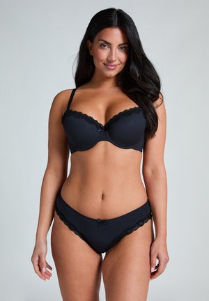 Schwarzer gerippter BH und passender Bikini mit Spitzenbesatz und einem kleinen Schleifendetail. Der BH hat Unterwäscheschalen und verstellbare Träger.