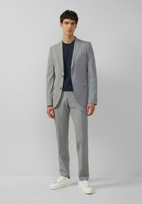 Lichtgrijze colbert en broek van gestructureerde stof, gecombineerd met een marineblauw t-shirt en witte sneakers, staande tegen een effen achtergrond.
