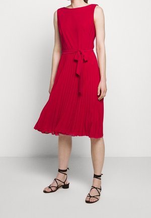 Rotes, ärmelloses Kleid mit einem fließenden, plissierten Rock und einer gebundenen Taille. Kombiniert mit schwarzen Riemchensandalen. Glatte Stofftextur.