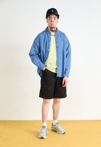 Camisa de denim, camiseta verde, pantalones cortos negros, zapatillas grises. Modelo de pie sobre un suelo amarillo con un fondo sencillo y una gorra negra.