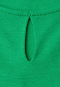 Tissu vert avec une ouverture verticale en forme de trou de serrure sous une couture horizontale cousue, montrant la texture et le motif tricoté.