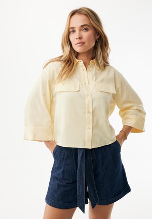 Lichtgeel overhemd met knopen en twee borstzakken, driekwart mouwen. Gecombineerd met donkere denim shorts, gestrikt in de taille met een riem.