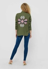 Camicia verde oliva con un design ricamato sul retro in rosa e bianco, abbinata a jeans skinny blu e tacchi beige.