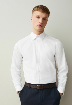 Young man with short blond hair wearing a white button-up shirt, brown belt, and dark pants, standing with hands in pockets.
