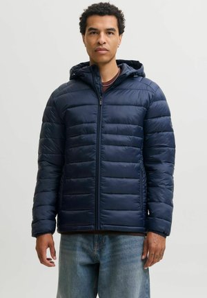 Homme portant une veste matelassée bleu marine à capuche avec fermeture éclair devant et un jean bleu clair sur un fond uni.