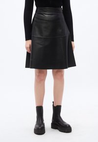 Μαύρη δερμάτινη φούστα σε γραμμή Α με ψηλή μέση, με απαλό υφή και εμφανείς ραφές. Συνδυάζεται με χοντρά μαύρα μποτάκια αστραγάλου.