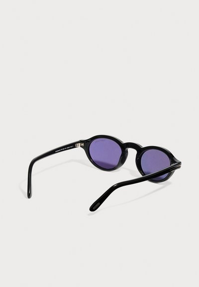 Lunettes de soleil rondes noires avec des verres violets, posées ouvertes sur une surface blanche, vues de dos et de côté.