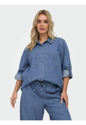 Blonde vrouw die een blauw gestreept denim overhemd met opgerolde mouwen en bijpassende broek met trekkoord draagt, tegen een effen achtergrond.