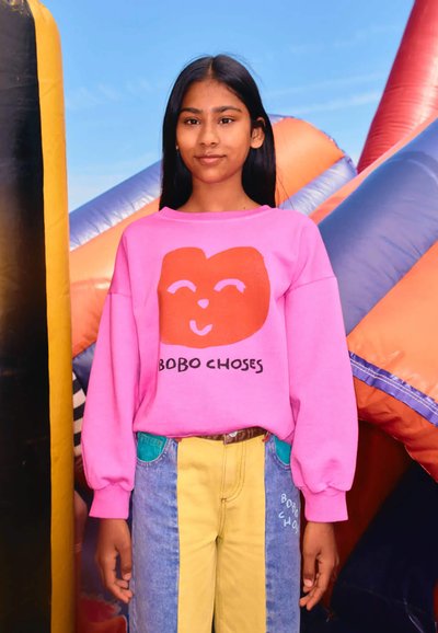 Pink sweatshirt with a red bear face graphic and text "BOBO CHOSE." Blue and yellow paneled jeans with pockets. Smooth fabric and relaxed fit.