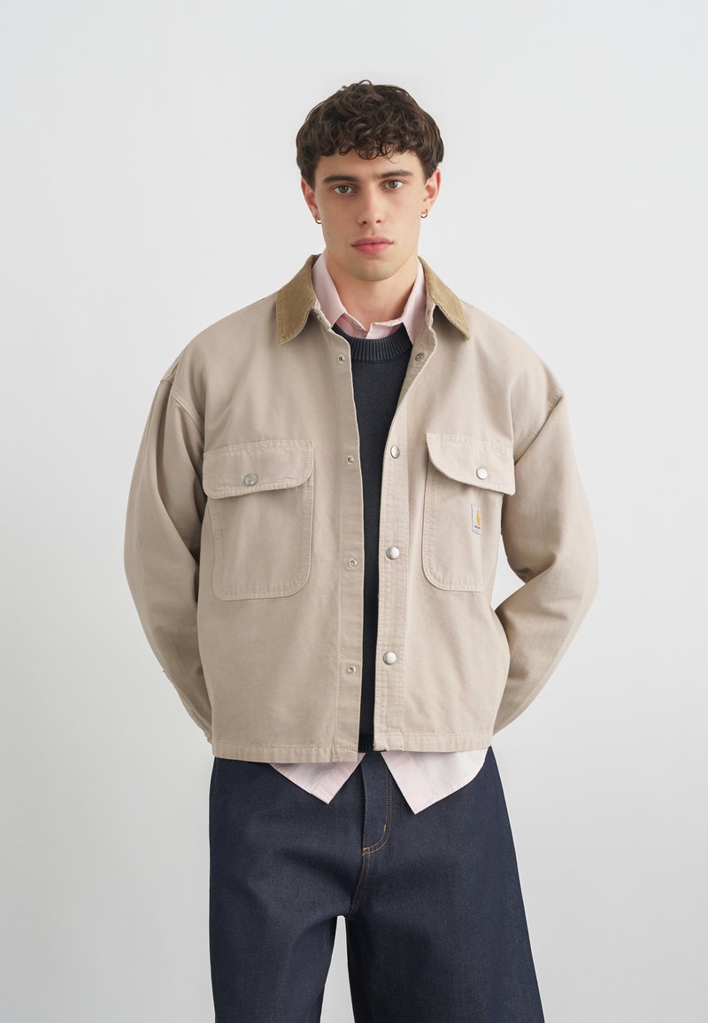 Joven con cabello corto y rizado, lleva una chaqueta beige abotonada sobre un suéter negro y una camisa rosa claro, de pie frente a un fondo liso.