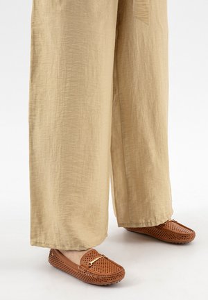 Pantalon beige à jambes larges porté avec des mocassins en cuir marron perforé, ornés d'un petit détail en barre d'or sur le dessus.