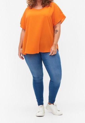 Orange kortärmad topp med avslappnad passform, tillverkad av textilmaterial. Kombinerad med blå jeans och vita sneakers, med en enkel design.