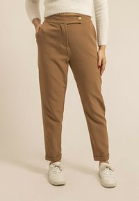 Pantalons marron ajustés avec une taille haute, des poches avant et un bouton en or à la taille. Associés à des baskets blanches. Texture lisse.