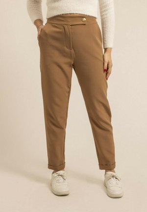 Pantalons marron ajustés avec une taille haute, des poches avant et un bouton en or à la taille. Associés à des baskets blanches. Texture lisse.
