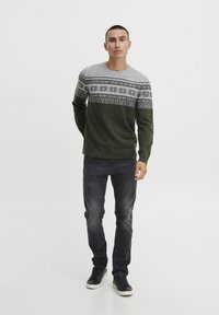 Grøn og grå strikket sweater med mønstrede skuldre, suppleret med mørke jeans og sorte sneakers. Modellen står foran en ensfarvet baggrund.