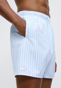 Shorts de bain bleu clair avec rayures blanches verticales, taille élastique, poches latérales et petit logo blanc sur le côté inférieur gauche.