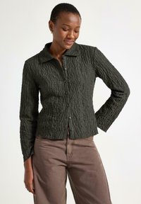 Mørkegrøn tekstureret skjorte med krave, lange ærmer og knaplukninger, parret med lysebrune bukser. Bemærkelsesværdig krøllet stofdetalje.