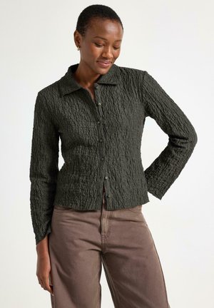 Tmavě zelená textura košile s límečkem, dlouhými rukávy a knoflíkovým zapínáním, v kombinaci se světle hnědými kalhotami. Výrazný detail zkrčené látky.