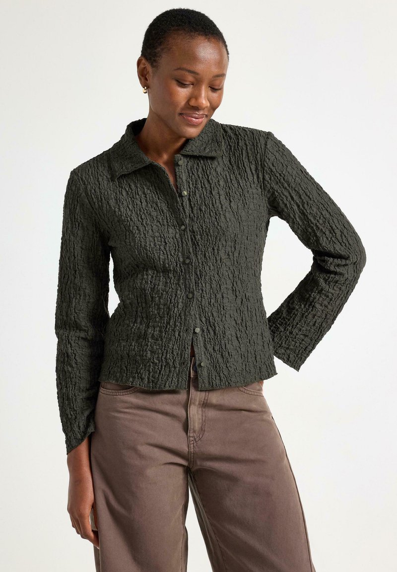 Mørkegrøn tekstureret skjorte med krave, lange ærmer og knaplukninger, parret med lysebrune bukser. Bemærkelsesværdig krøllet stofdetalje.