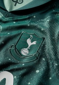 Maglia sportiva verde in tessuto con un design texturizzato, un logo con un uccello e un pallone da football, e un colletto a v. Le rifiniture includono dettagli bianchi.