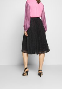 Mustat laskostetut hameet kiiltävät, yhdistettynä vaaleanpunaiseen paitapuseroon, jossa on kuvioidut hihat. Mustat avonaiset korkokengät korostavat asua.