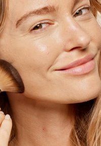 Makeupborste med en avsmalnande handtag som applicerar produkt på en kind. Huden ser slät ut med en naturlig textur. Håret är vågigt och löst.
