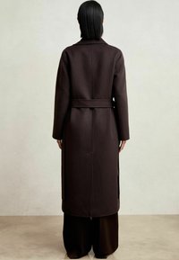 Garšs, tumši brūns vilnas mētelis ar jostu, ar izgrieztiem apkakli un sānu šķēlumiem. Audums izskatās strukturēts un ar tekstūru.