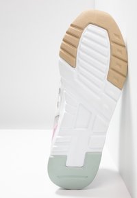 Semelle d'une basket montrant une bande de roulement blanche, beige et vert clair avec des motifs texturés, positionnée verticalement contre une surface blanche.