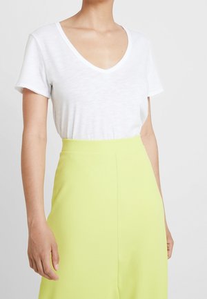 Vrouw draagt een witte, korte mouw, V-hals T-shirt, ingestopt in een hooggesneden felgele rok tegen een effen achtergrond.