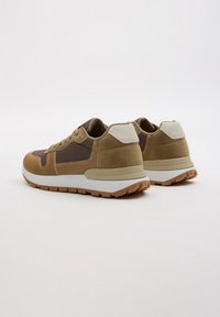 Zapatillas deportivas marrones y marrón claro con un upper texturizado, detalles en blanco y beige, y una suela de goma. Presenta cordones y un collar acolchado.