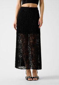 Gonna plissettata in pizzo nero con uno strato trasparente. Presenta una cintura arricciata e motivi floreali intricati. Abbinata a tacchi neri con fiocchi decorati.