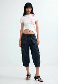 Joven mujer con una camiseta blanca recortada con ribete rosa y pantalones capri cargo negros, de pie descalza frente a un fondo liso.