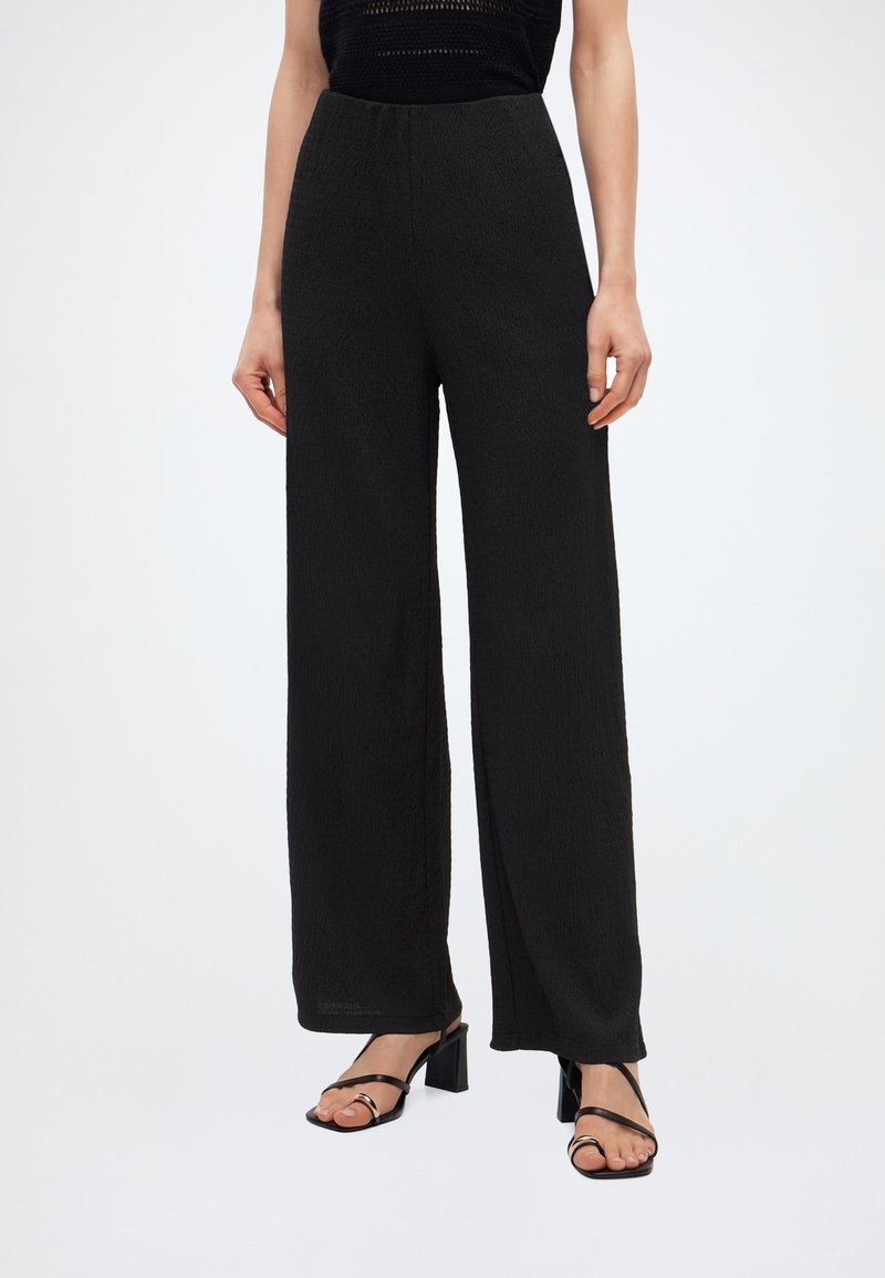 Γυναίκα που φοράει μαύρο παντελόνι ψηλόμεσο με φαρδιά μπατζάκια, συνδυασμένο με μαύρα σανδάλια με τακούνι, λεπτές λουρίδες και χρυσούς κρίκους.