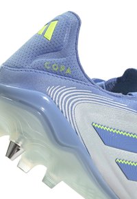 Chaussure de sport bleue avec une tige en tissu texturé, des accents argentés et une semelle en caoutchouc clair dotée de pointes pour une meilleure traction. Logo "COPA" visible.