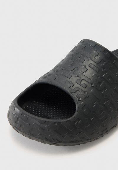 Ciabatta in gomma nera con punta aperta. Superficie testurizzata che presenta un motivo geometrico. L'interno include piccole punte rialzate per una migliore aderenza.