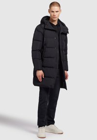 Fekete párnázott parkával, kapucnival, gombos elülső résszel, oldalsó zsebekkel és középhosszú kialakítással, sötét farmerrel és fehér tornacipővel párosítva.