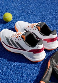 Witte sport schoenen met rode en zwarte accenten, een gestructureerde mesh bovenkant, oranje zool en contrasterende veters op een blauwe gestructureerde ondergrond.