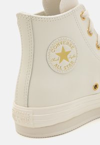 Weißes Leder-Hochschnitt-Sneaker mit einem runden Patch, das einen goldenen Stern und den Text "Converse All Star" zeigt. Goldene Hardware-Akzente, strukturierte Oberfläche.