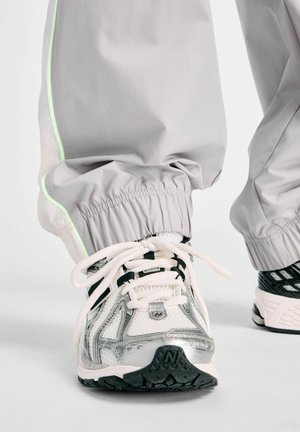 Nahaufnahme von weißen und silbernen Sportschuhen mit schwarzen Sohlen, getragen zu hellgrauen Hosen mit einem neongrünen Streifen nahe den elastischen Bündchen.