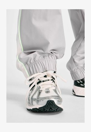 Close-up van witte en zilveren sportschoenen met zwarte zolen, gedragen met lichtgrijze broek met een neon groene streep bij de elastische boorden.