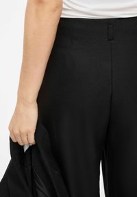 Črne hlače z teksturirano površino, ki imajo diskreten zadnji žep in oprijet pas. Roka drži črno jakno.